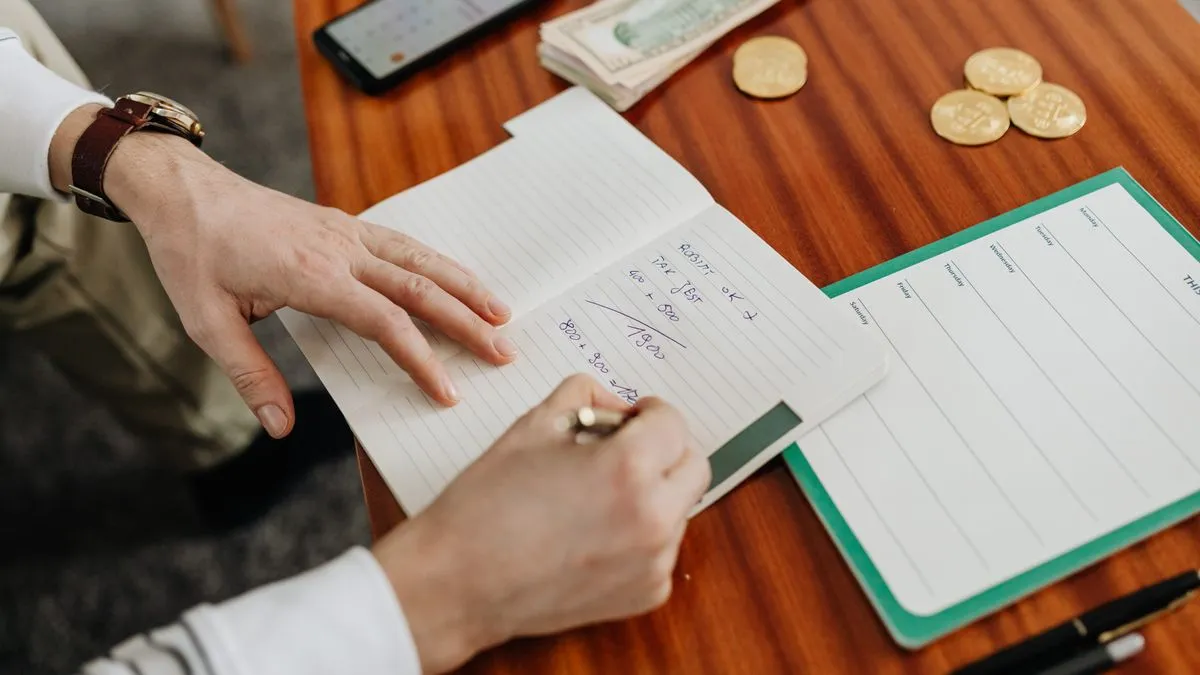 Emergency Fund: How Many Months Do You Really Need? A man's hands work through handwritten sums in a lined notebook, with folded banknotes, a few gold coins and a weekly planner sheet on the desk beside him.