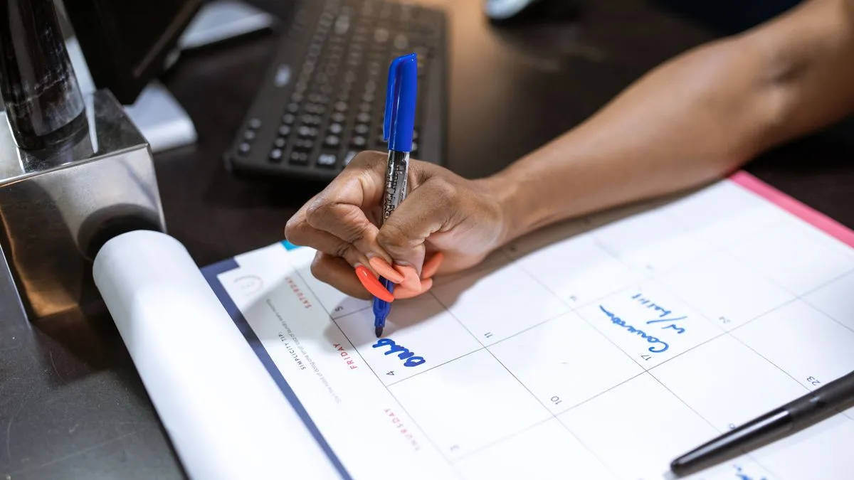 ETF savings plan: how to automate your investing A person at an office desk writes an entry into a paper desk planner with a blue ballpoint pen, a computer keyboard behind it.