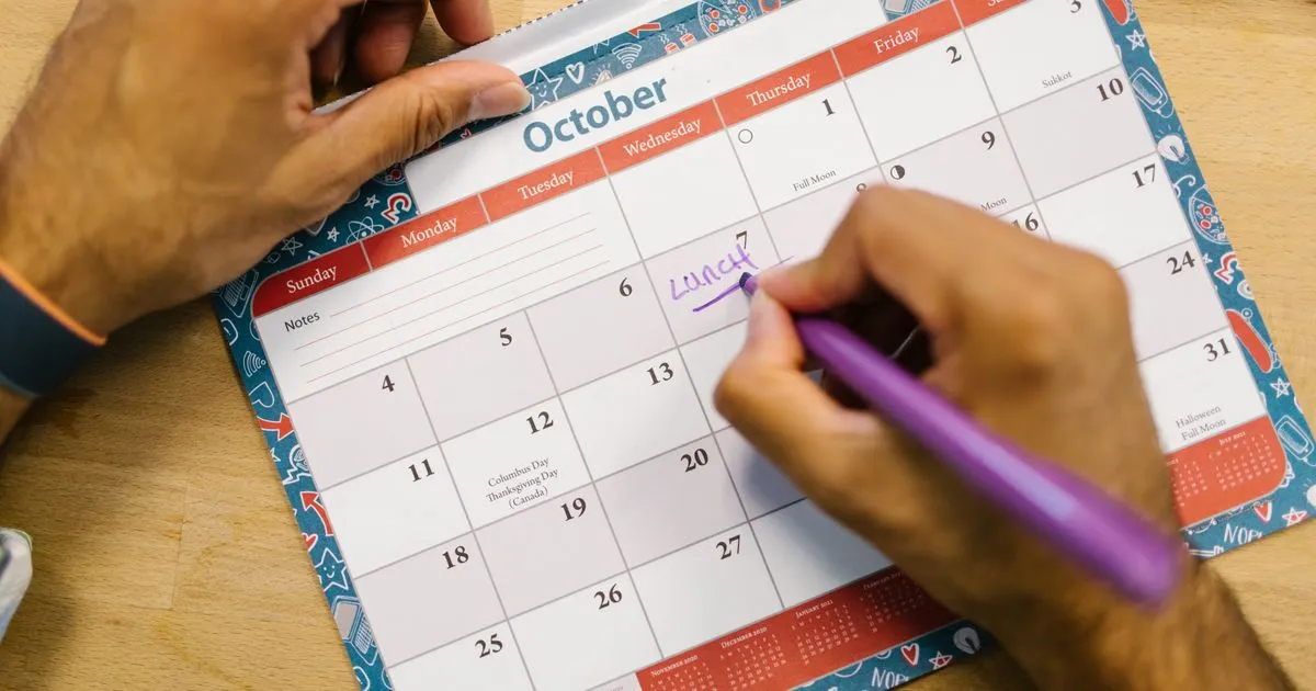 Two hands mark a date with a purple pen on a paper wall calendar open to the month of October.