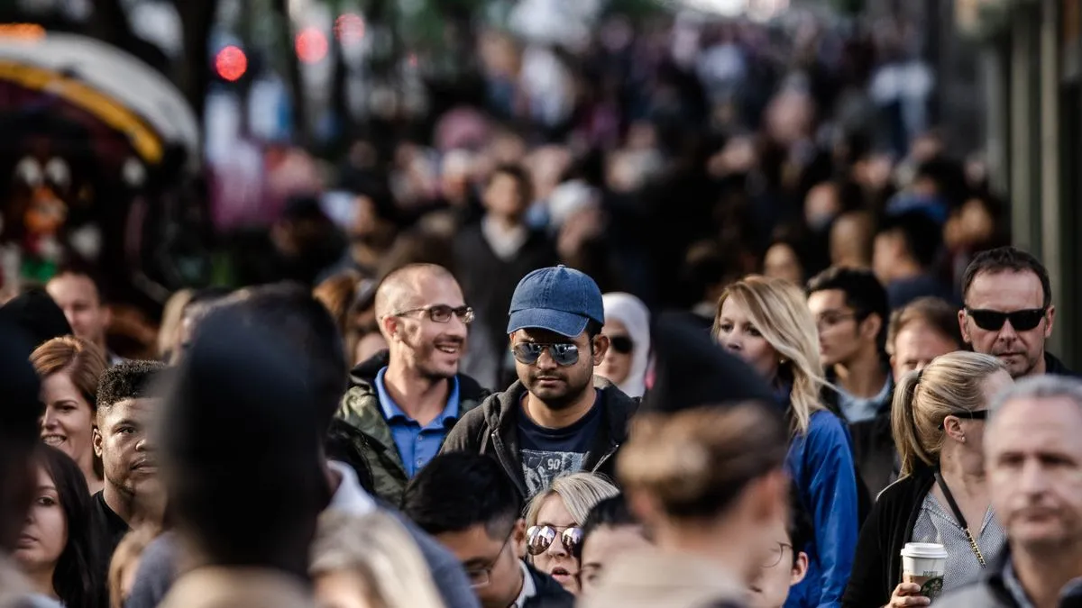 Net worth percentile by age: how to read the data A close-up of a crowded street, one man in a blue cap and sunglasses in sharp focus among dozens of blurred pedestrians.