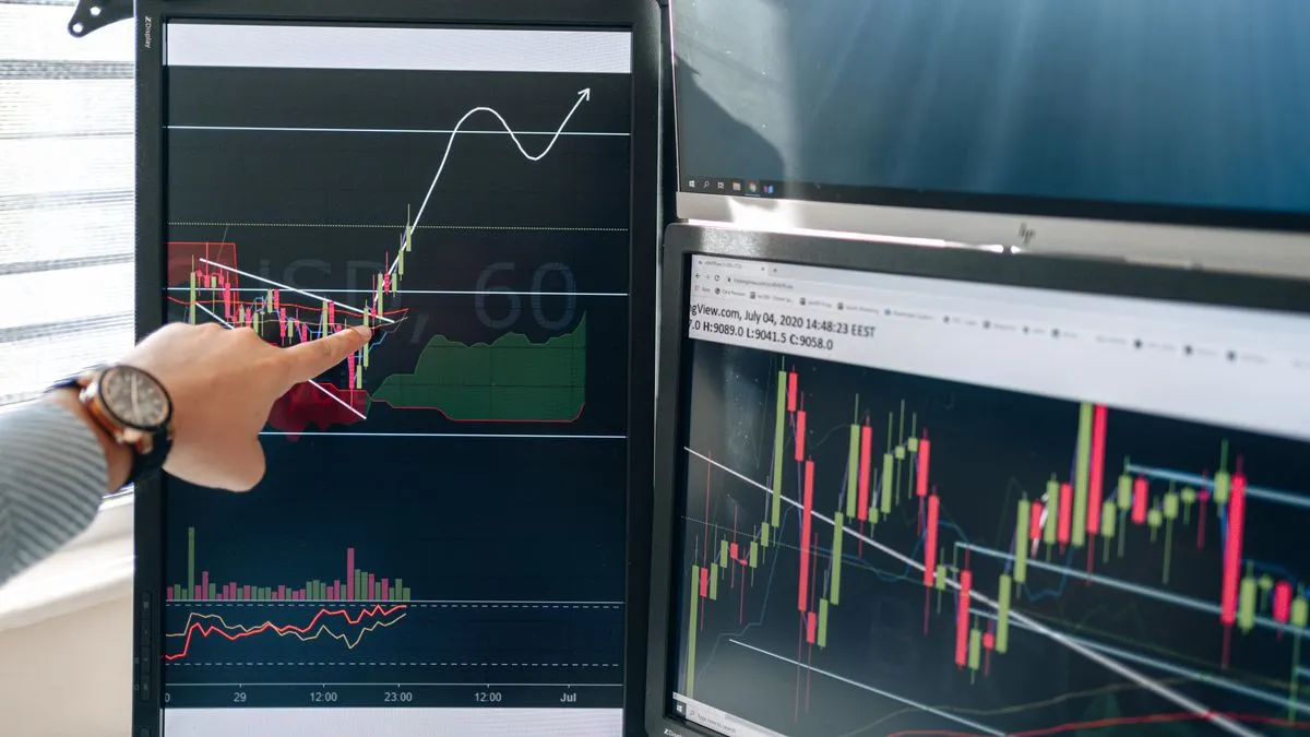 Option Greeks Explained: Delta, Gamma, Theta, Vega A hand with a wristwatch points at an upward arrow sketched over a candlestick chart on a vertical monitor, with more charts on a screen behind.