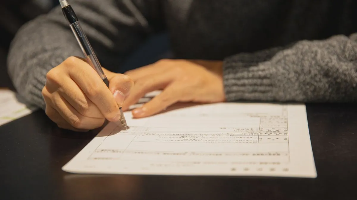 Freistellungsauftrag: Split the German Savers Allowance A person in a grey sweater fills in a printed form with a ballpoint pen at a dark wooden table.