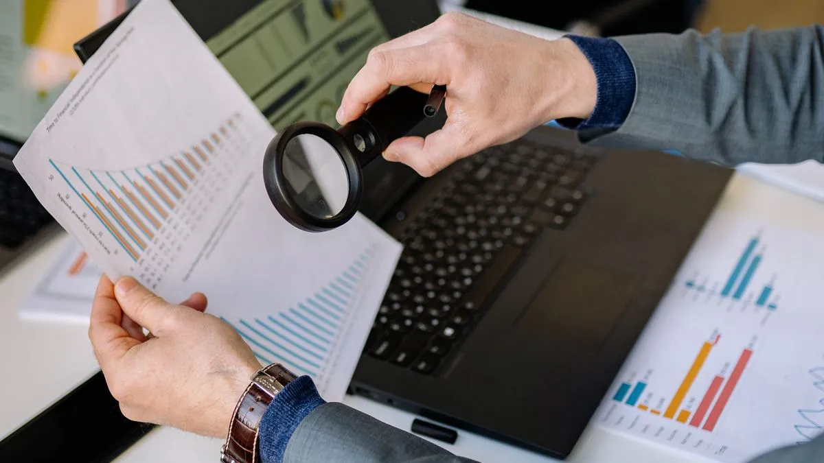Valuation Metrics: How to Tell If a Stock Is Cheap Hands hold a magnifying glass over printed pages of bar charts and figures next to a laptop.